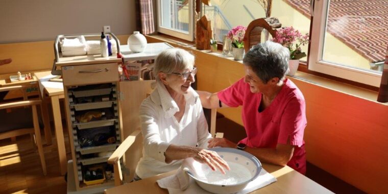 Pflegekraft hilft älterer Dame beim Entspannen mit einer Handmassage in einem hellen, freundlichen Raum.